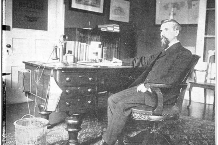 Black-and-white photograph of a late 19th-century man seated at a desk in a Wall Street–style office, surrounded by books, papers, and period furnishings.