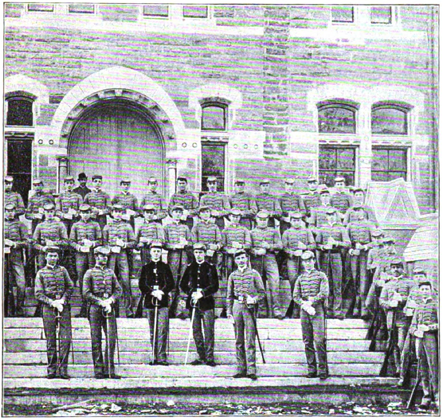 Military cadets in uniform arranged in rows before stone university building