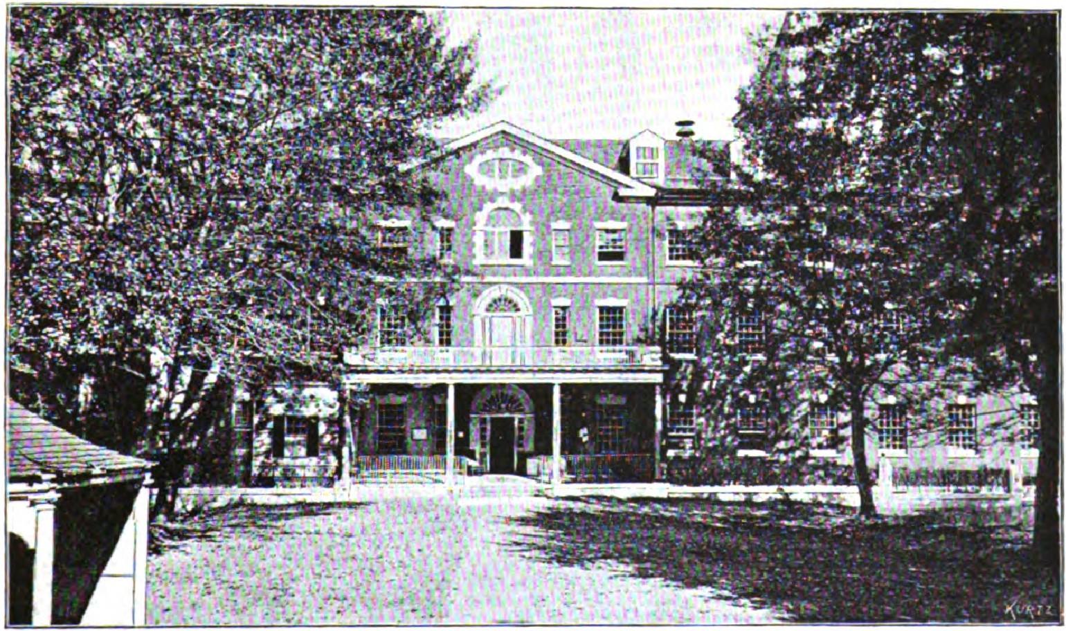 Historic colonial-style building with front porch surrounded by large trees